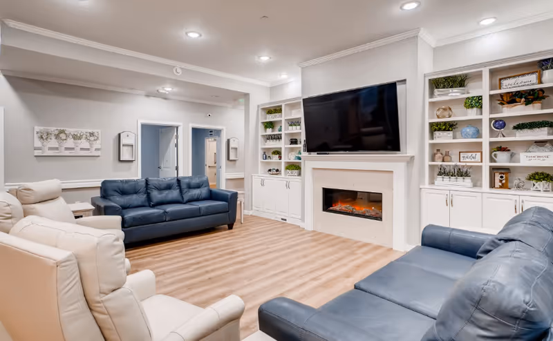 Bright communal living room with leather sofas, a fireplace, wall-mounted TV and built-in shelving filled with decor.