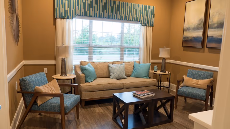 Bright living room with a beige sofa, two blue patterned armchairs, a dark coffee table, lamps and a large window.