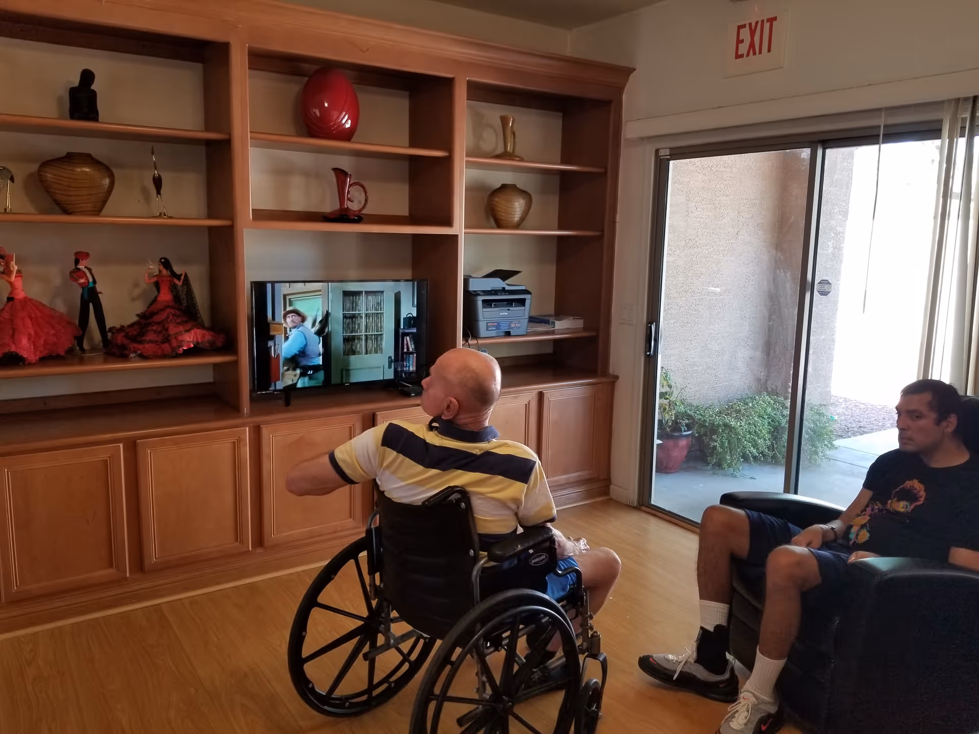 Two men, one in a wheelchair, watching television in a common living room with built-in wooden shelving and a sliding glass door.