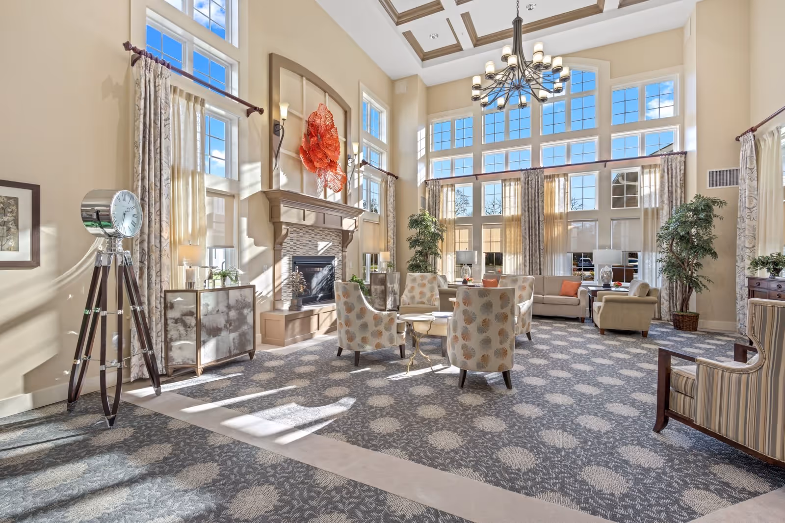 A spacious and well-lit living room area with large windows allowing natural light to fill the space. The room features a high ceiling with a decorative chandelier, a fireplace with a large red decorative piece above it, several upholstered chairs with patterned fabric, a sofa, armchairs, potted plants, and a large tripod floor clock. The carpet has a floral pattern, and curtains frame the windows.