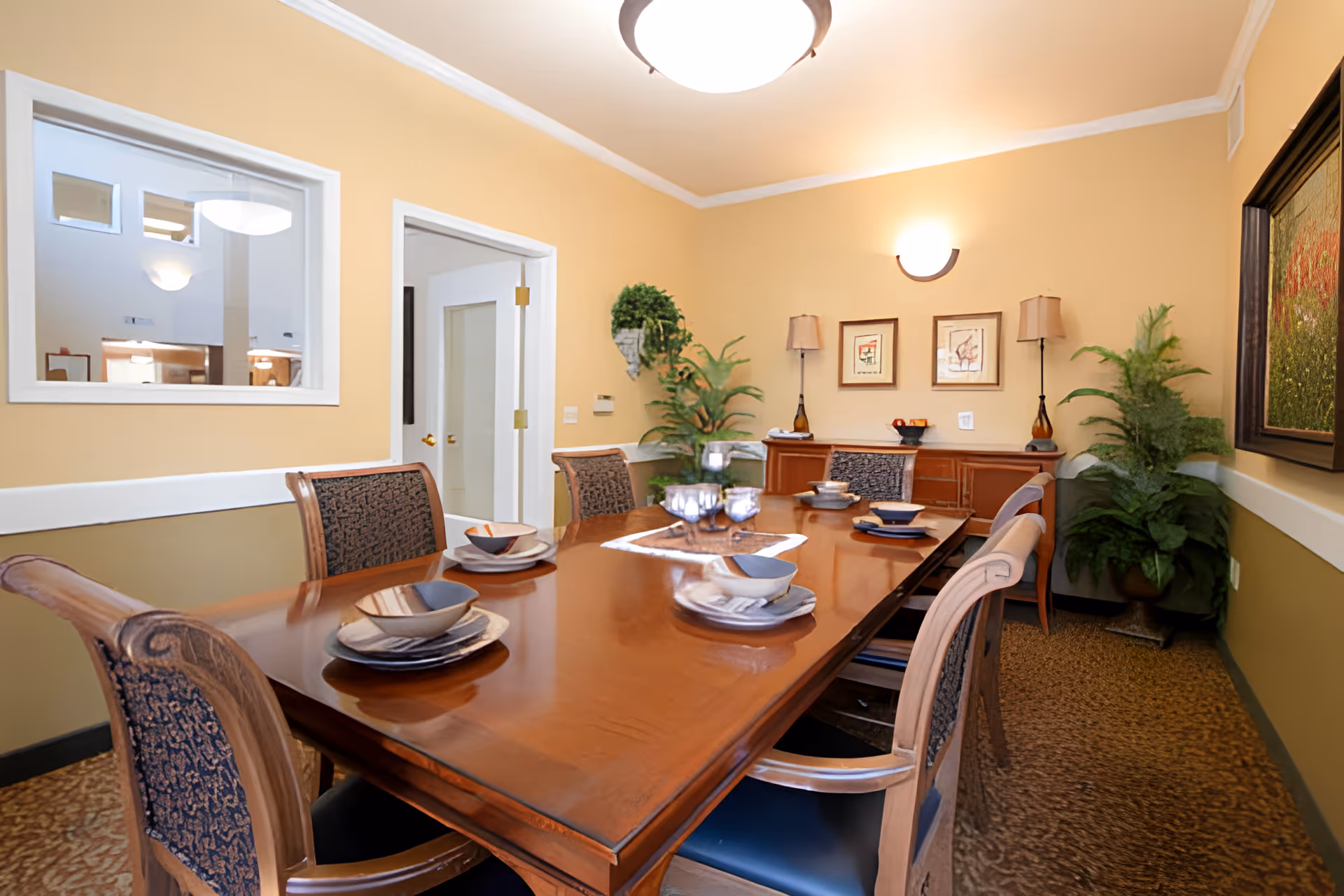 A dining room with a long wooden table set with plates, bowls, and glasses. The room has beige walls with white trim, a sideboard with two lamps and framed artwork above it, and two potted plants in the corners. There is a window and a door on one wall, and a ceiling light fixture.