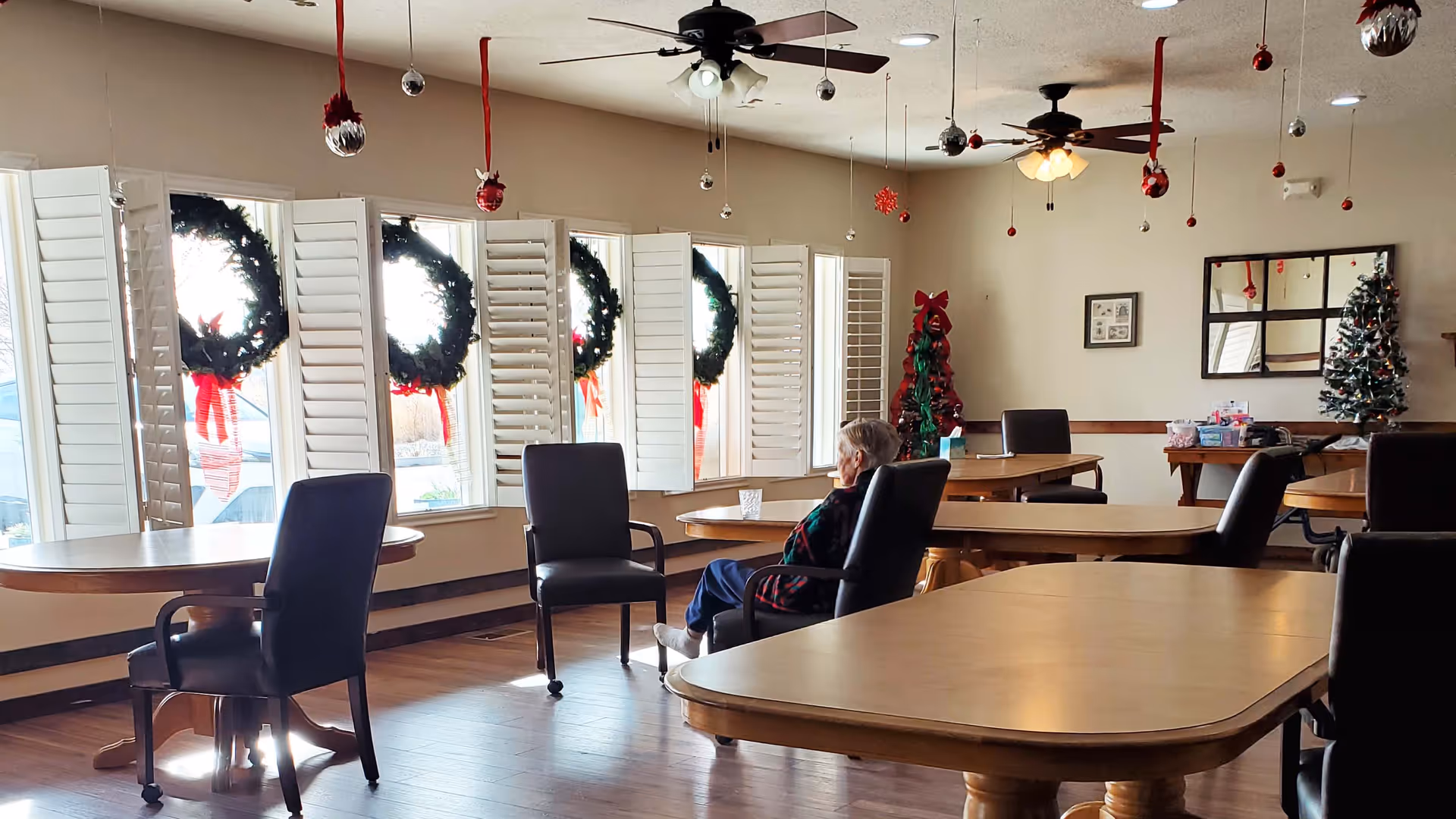 An elderly resident sits at a table in a holiday-decorated communal dining room with wreaths on the windows.