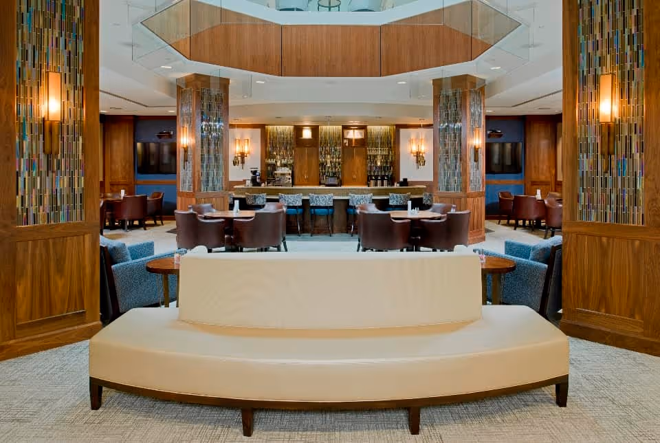A spacious and elegant common area with a curved beige sofa in the foreground, surrounded by wooden pillars with decorative glass panels and wall sconces. In the background, there are several round tables with brown leather chairs and a bar area with high stools and a mirrored backdrop.