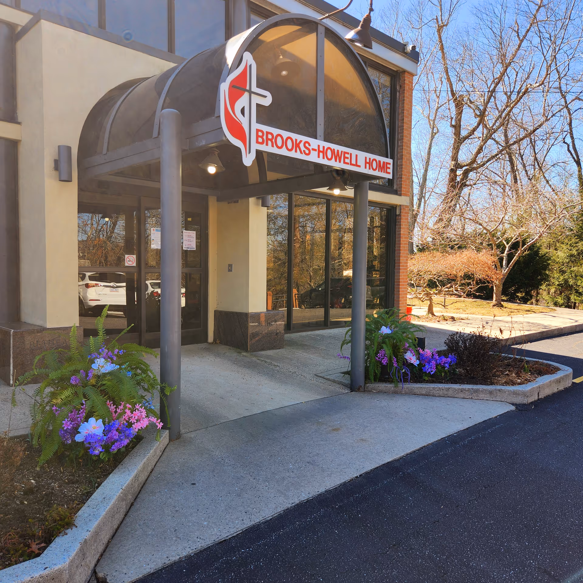 Entrance to Brooks-Howell Home with a covered walkway supported by metal poles. There are flower beds with purple and pink flowers on either side of the entrance. The building has large glass windows and a sign above the entrance displaying the facility's name.
