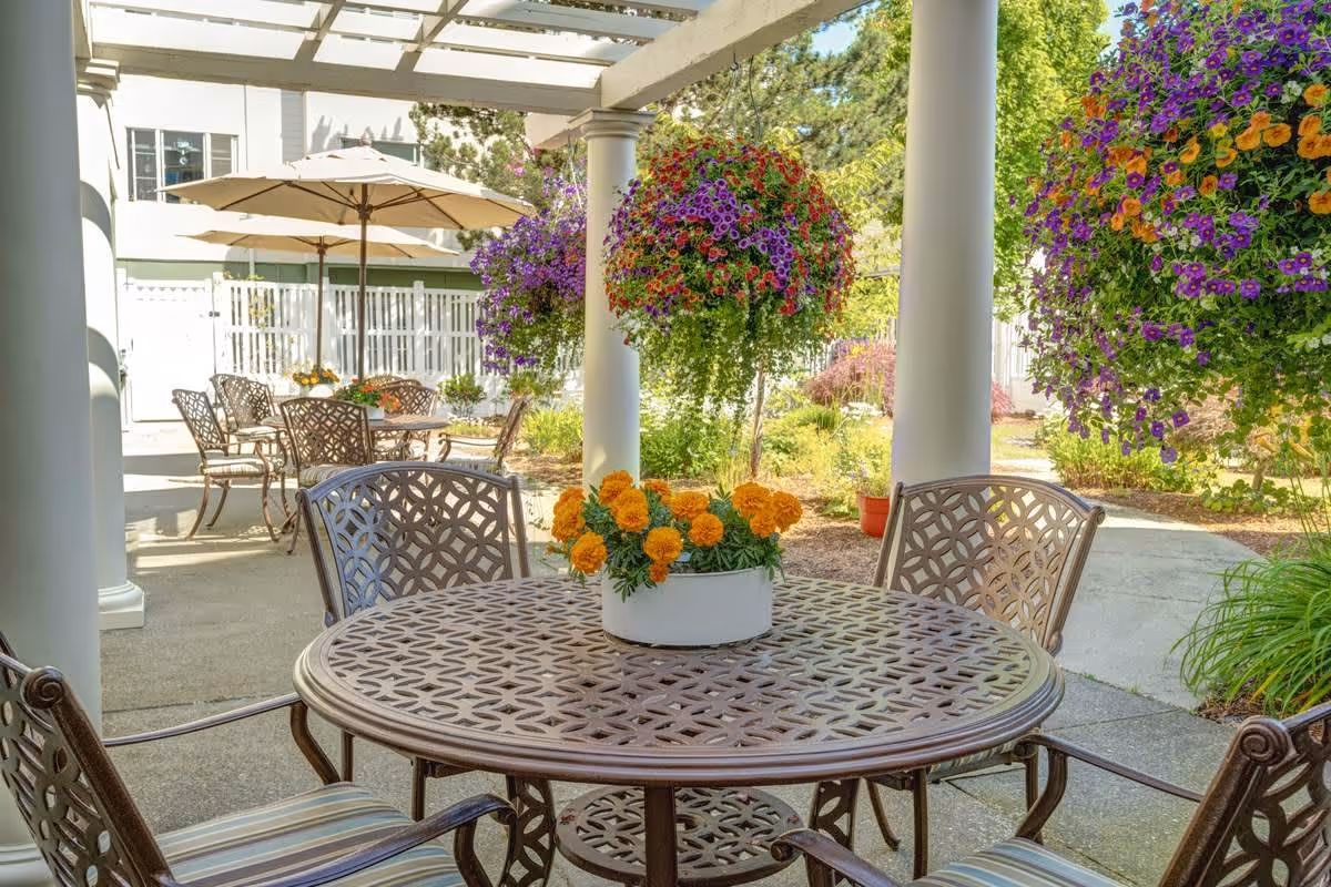 Outdoor patio area with metal tables and chairs, some tables have umbrellas. Hanging baskets with colorful flowers and a potted plant with orange flowers on the table. White columns and a pergola overhead, with greenery and a white fence in the background.