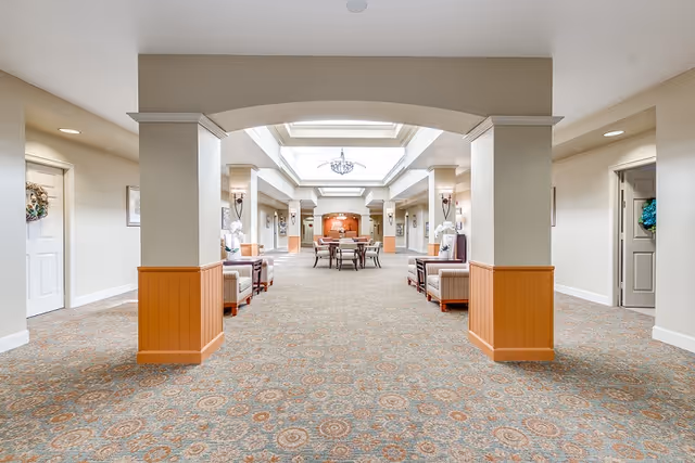 A spacious, well-lit common area in a senior living facility with patterned carpet, beige walls, and wooden paneling on the lower half of the columns. The room features comfortable armchairs arranged along the sides and a table with chairs in the center under a skylight with a chandelier. Doors and decorative wreaths are visible along the walls.