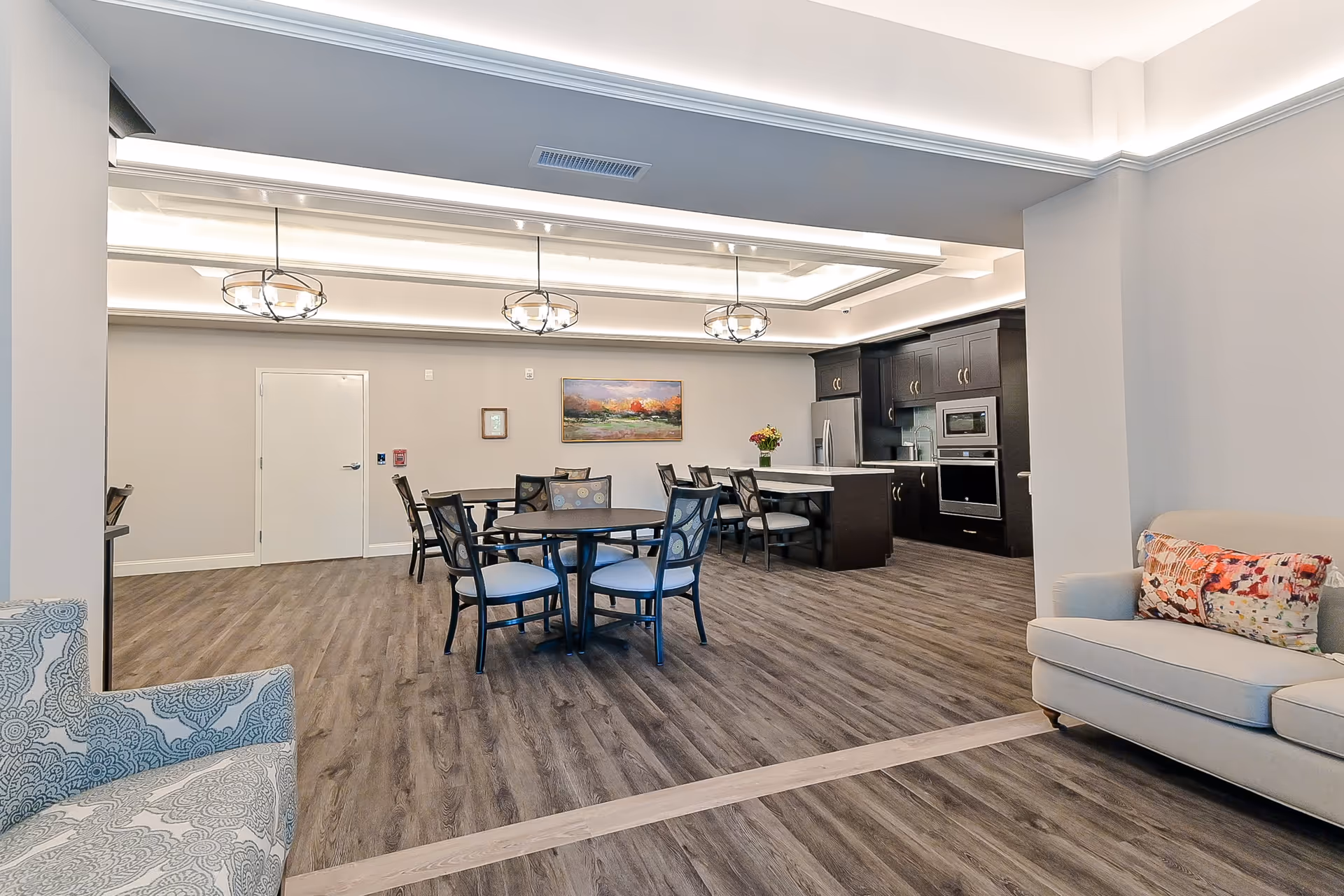 A modern common area with a kitchen and dining space. The room features dark wood cabinets, stainless steel appliances, a kitchen island with chairs, round dining tables with chairs, and comfortable seating including a patterned armchair and a beige sofa with a colorful pillow. The flooring is wood-style, and the ceiling has recessed lighting and three decorative pendant lights.