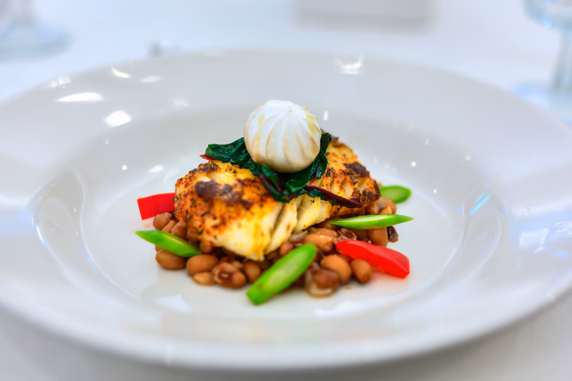 A plated gourmet dish featuring a piece of grilled fish on a bed of beans, garnished with green snap peas, red bell pepper slices, leafy greens, and topped with a small dollop of white sauce or cream, served on a white plate.