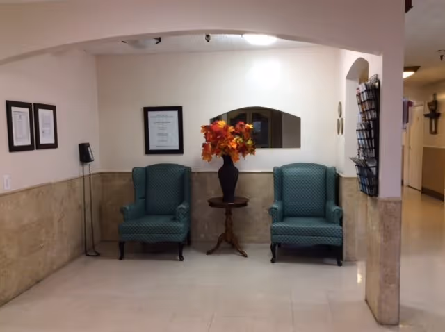 A small seating area with two green upholstered armchairs separated by a wooden side table holding a vase with orange and red artificial flowers. The walls are beige with a tiled lower half, and there are framed certificates or documents on the left wall. A small window-like opening is above the table, and a brochure rack is mounted on the right wall near an archway leading to a hallway.
