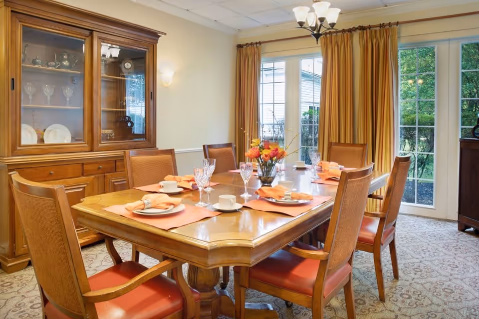 A dining room with a wooden table set for six people, featuring orange placemats, napkins, white cups, and crystal glasses. There is a flower arrangement in the center of the table. The room has a wooden china cabinet with glass doors displaying plates and glassware, beige walls, patterned carpet, and large windows with yellow curtains letting in natural light.