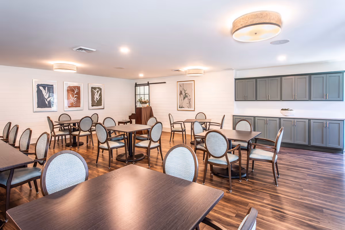A bright and spacious dining room with multiple wooden tables and cushioned chairs arranged neatly. The room features wooden flooring, white shiplap walls adorned with framed botanical artwork, and gray cabinetry along one wall. Ceiling lights provide warm illumination, and a piano is visible in the corner near a decorative window panel.