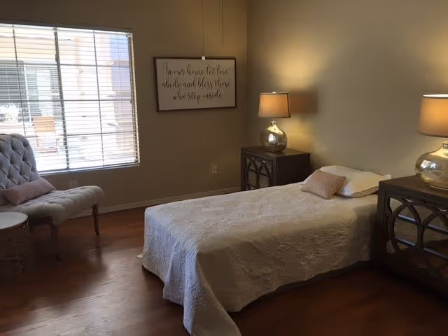 A bedroom with a single bed covered with a white quilt and a pillow. There are two matching bedside tables with lamps on each side of the bed. A cushioned chair with a pink pillow is placed near a window with blinds. A framed sign with cursive writing hangs on the wall above the bed.