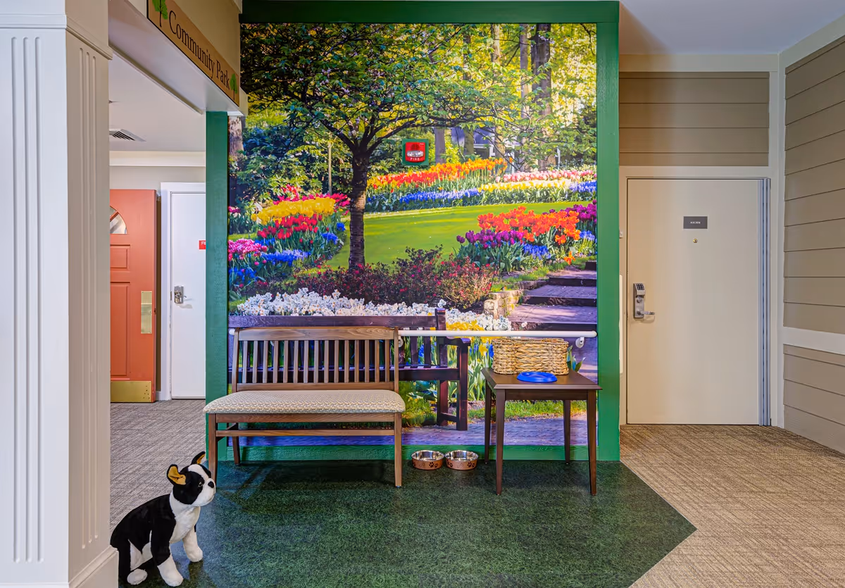 Indoor seating area with a wooden bench and a small table holding a basket and a blue frisbee. Behind the bench is a large wall mural depicting a colorful garden with flowers and a tree. On the floor in front of the bench are two pet bowls. To the left, there is a stuffed dog toy near a white column. The area has green and beige flooring, and there are doors visible in the background, one labeled 'KITCHEN'.