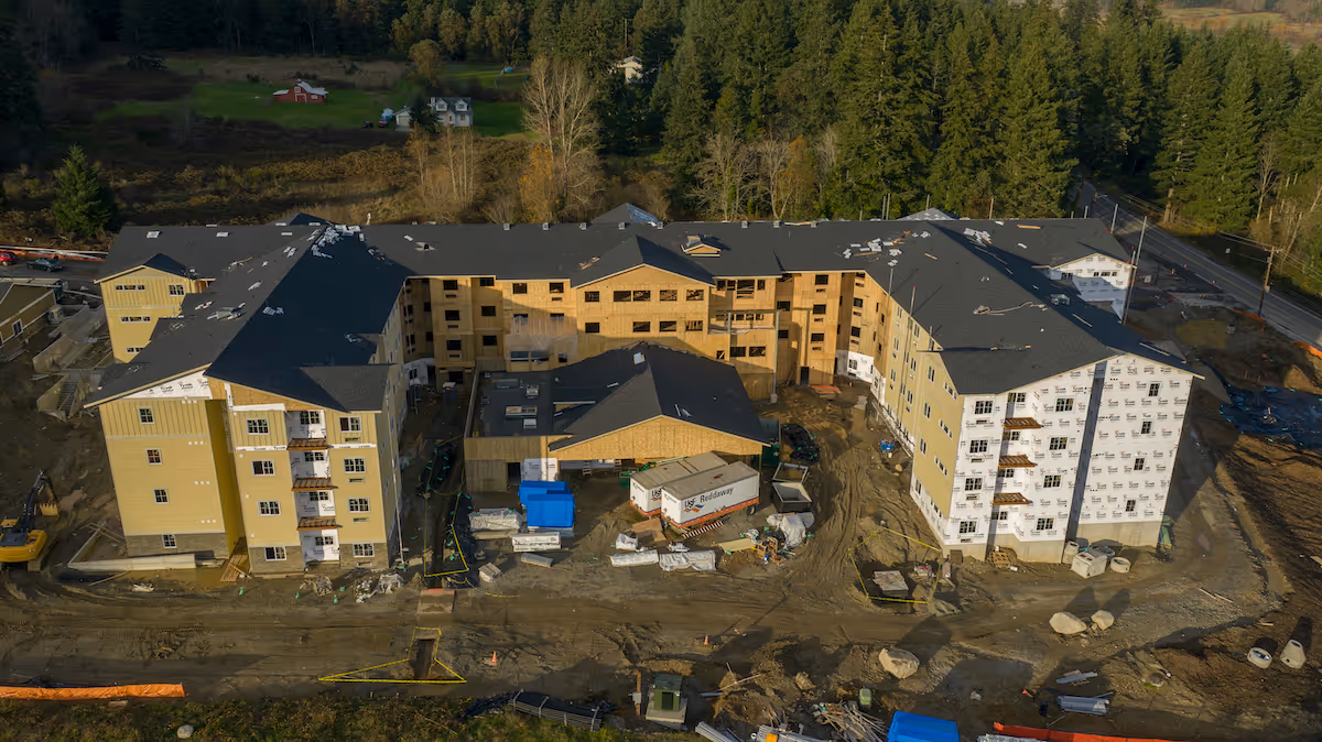 Aerial view of a large multi-story building under construction surrounded by dirt and construction materials, with a forested area in the background.