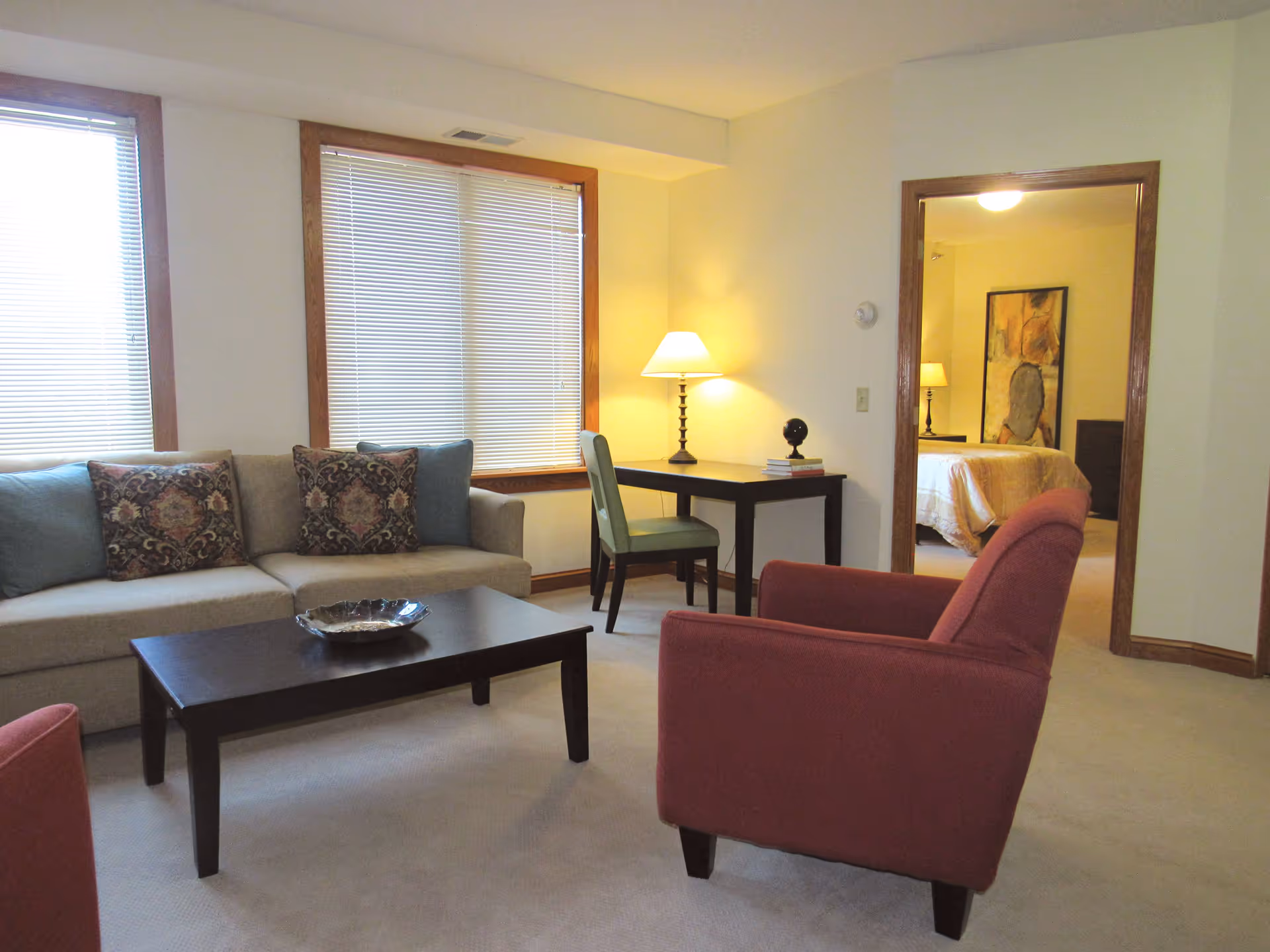 A cozy living room with a beige sofa adorned with patterned and solid pillows, a dark wooden coffee table with a decorative bowl, and a red armchair. There are two large windows with blinds, a small desk with a green chair and a lamp, and an open doorway leading to a bedroom with a bed, nightstand, lamp, and a framed artwork on the wall.