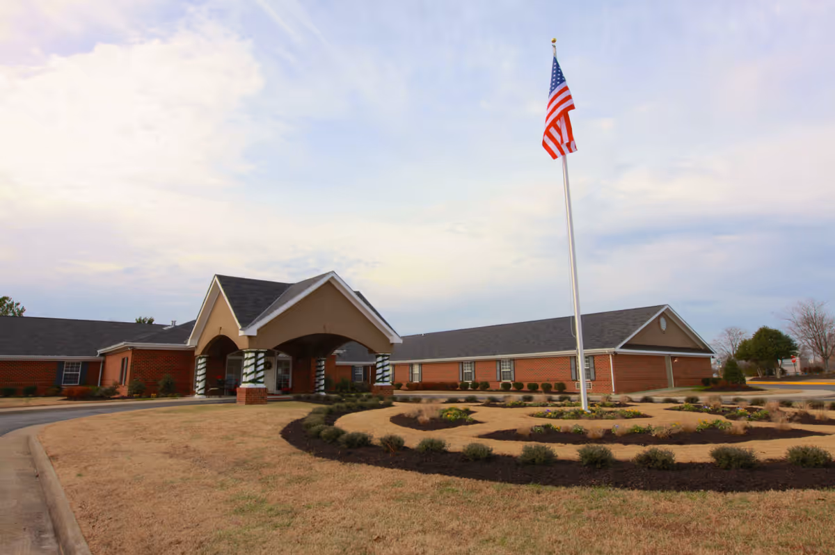 Exterior view of a single-story brick building with a covered entrance supported by columns wrapped in decorative material. In front of the building is a circular landscaped area with small bushes and an American flag on a tall flagpole. The sky is partly cloudy.