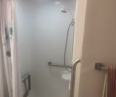 A small shower area with a white shower curtain featuring a cactus pattern, a handheld showerhead attached to the wall, a grab bar for support, and a towel hanging on a hook outside the shower.