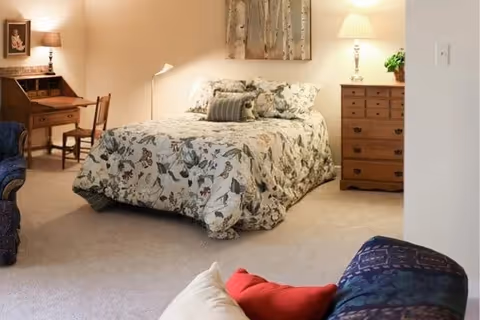 A cozy bedroom with a floral-patterned bedspread on a double bed, a wooden dresser with a lamp and plant on top, a wooden desk with a chair and lamp, and a comfortable seating area with colorful pillows in the foreground.