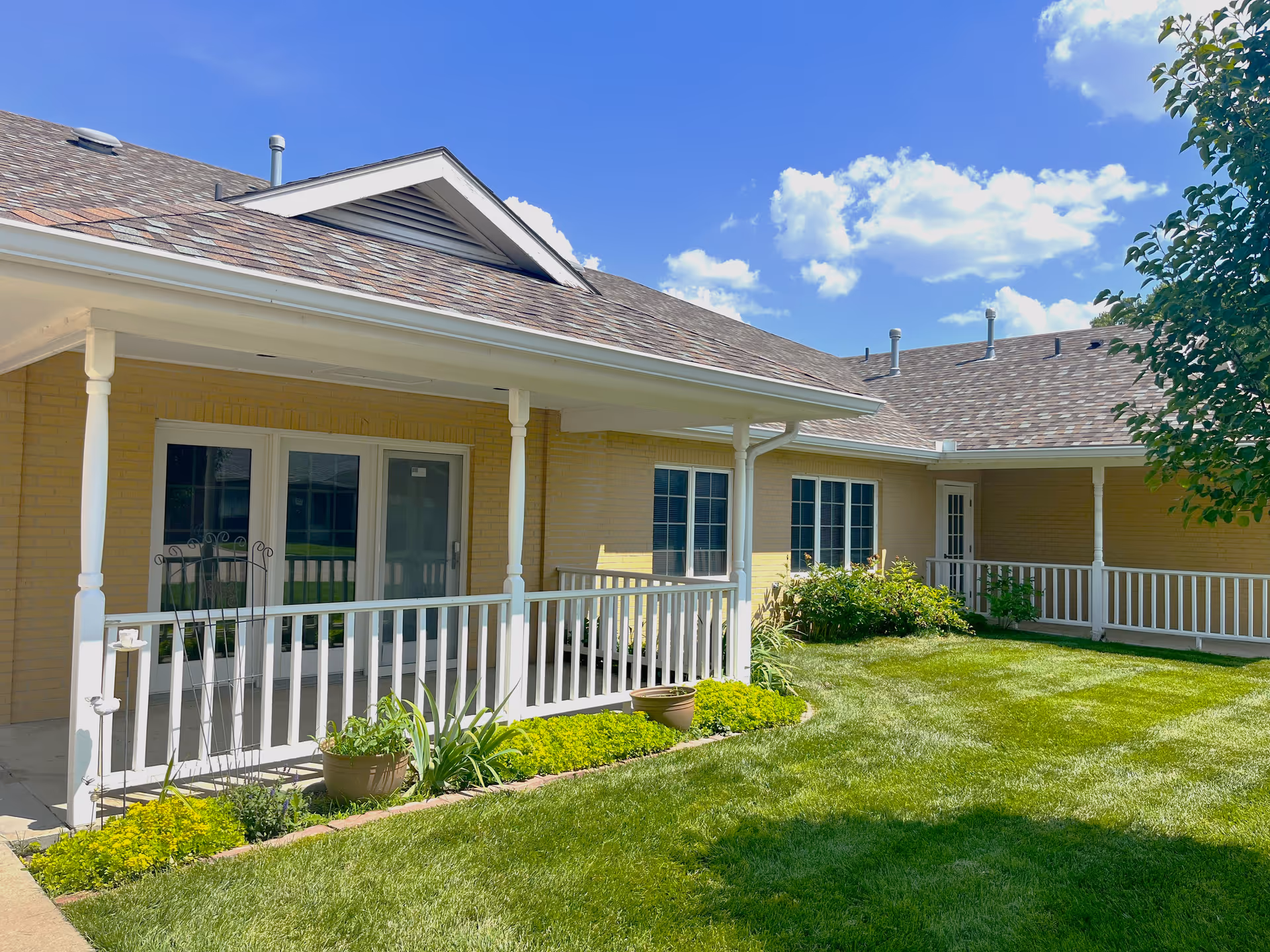 Covered porch with white railing and potted plants overlooking a green lawn in front of a single-story brick building under a blue sky.