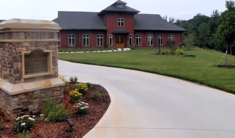 A large red building with multiple windows and a dark roof, situated at the end of a curved concrete driveway. In the foreground, there is a stone monument sign with a plaque and landscaped flower beds. The surrounding area has green grass and trees in the background.