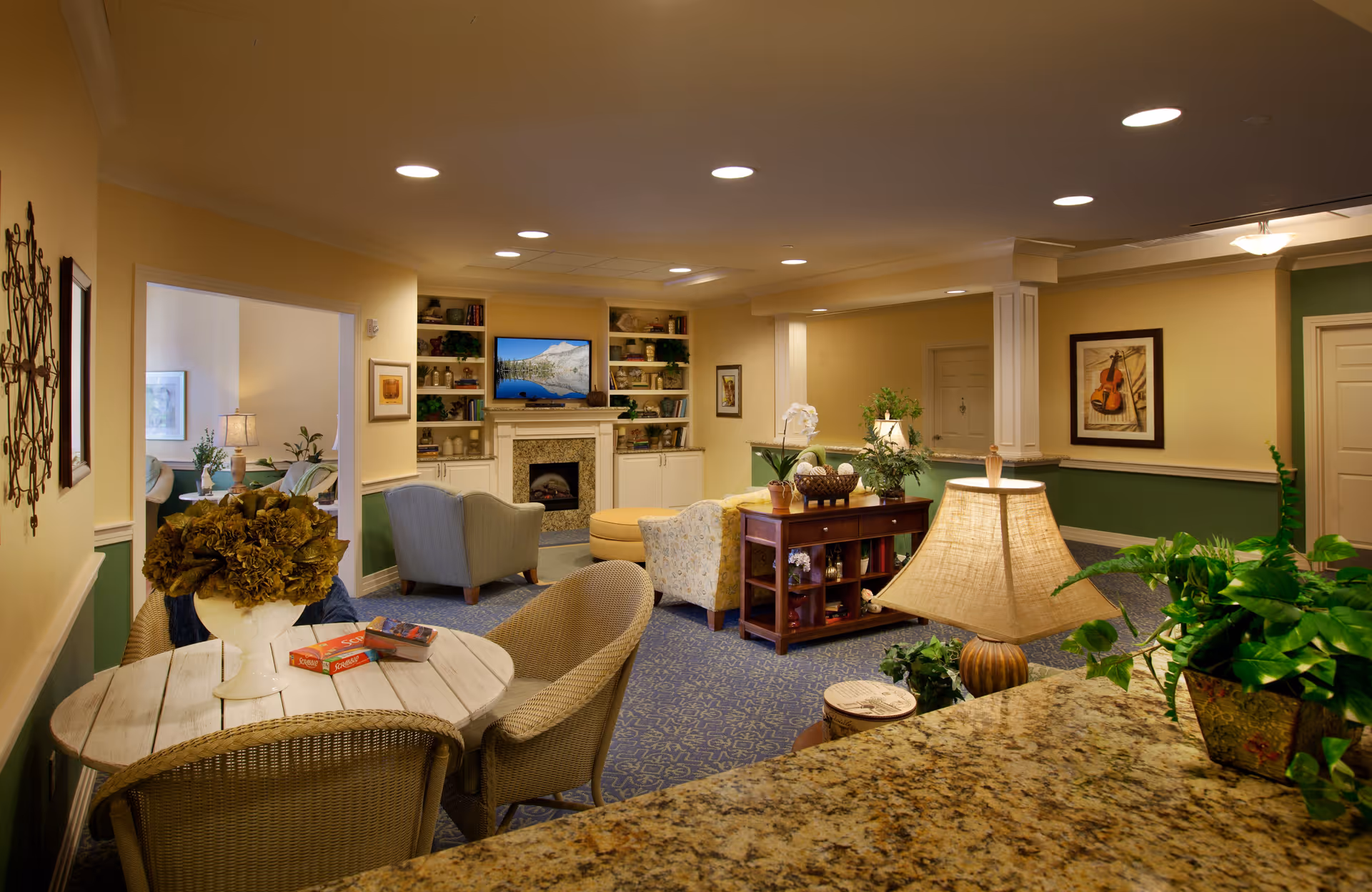 A cozy senior living facility common area with comfortable armchairs, a round table with wicker chairs, a lamp on a granite countertop, and various plants. The room features a fireplace with a TV above it, built-in shelves with decorative items, and framed artwork on the walls. The color scheme includes warm yellow walls with green wainscoting and a patterned blue carpet.