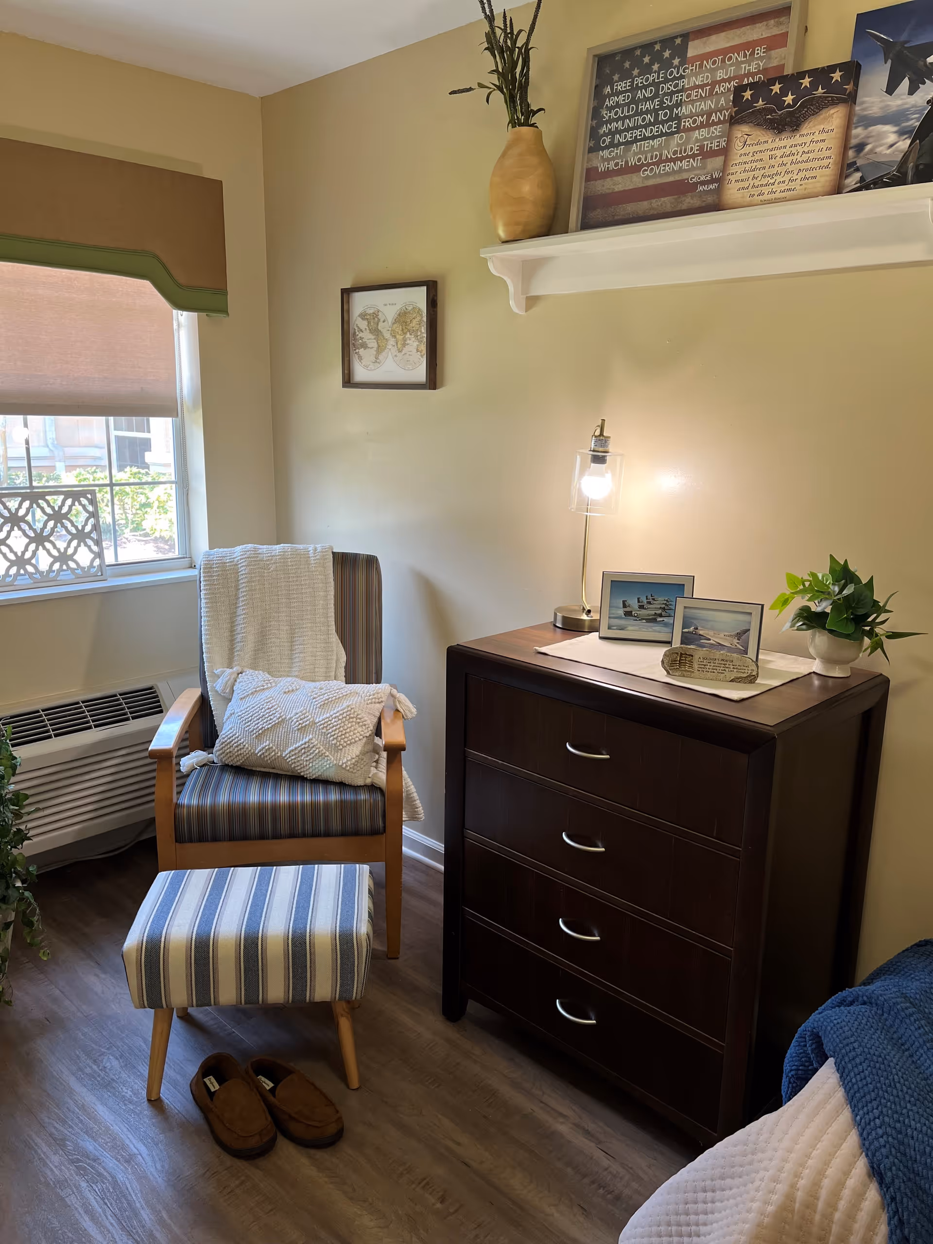 A cozy corner of a room featuring a striped armchair with a white textured pillow and a white throw blanket draped over it. In front of the chair is a matching striped footstool and a pair of brown slippers on the wooden floor. To the right is a dark wooden dresser with four drawers, topped with a small lamp, framed pictures, a small plant, and decorative items. A window with a beige shade and green valance lets in natural light. The walls are light beige with a small framed map and a shelf holding a vase and framed quotes.