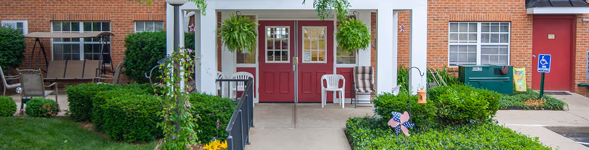 Front entrance of a brick senior living building with red double doors, a covered porch, hanging ferns, chairs, and surrounding shrubs.