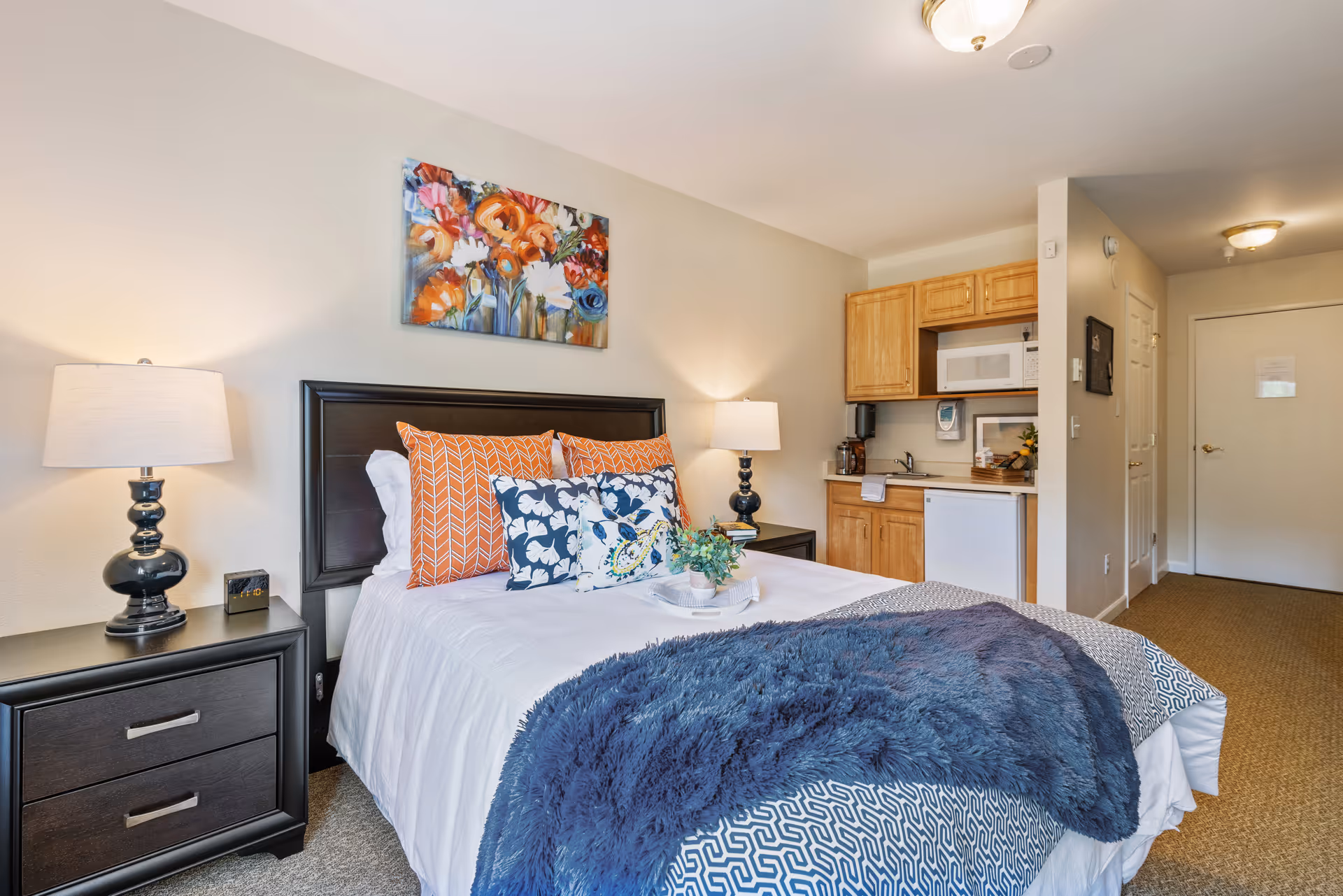 A cozy bedroom with a neatly made bed featuring white sheets, orange and blue patterned pillows, and a blue throw blanket. There are two bedside tables with matching lamps on either side of the bed. Above the bed hangs a colorful floral painting. To the right, there is a small kitchenette with wooden cabinets, a microwave, a sink, and a mini refrigerator. The room has beige walls and carpeted flooring, with a hallway leading to a closed door.
