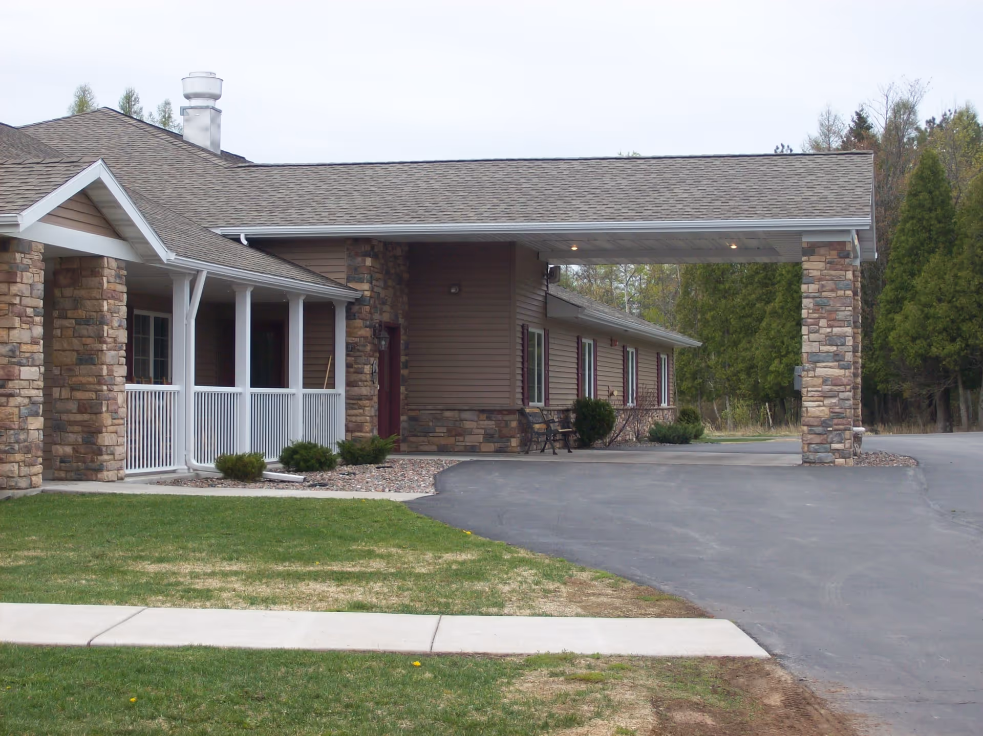 Exterior view of a senior living facility building with a covered entrance supported by stone pillars, a paved driveway, and a small lawn area with a sidewalk in front. The building has a combination of stone and siding on the walls and several windows along the side.