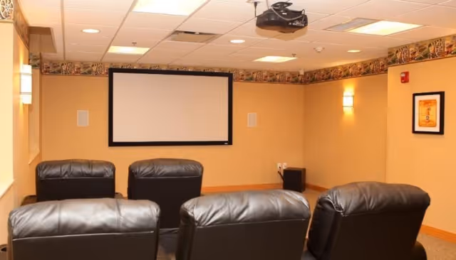 A small home theater room with four black leather recliner chairs facing a blank projector screen mounted on a beige wall. The room has a ceiling-mounted projector, recessed lighting, wall sconces, and decorative wallpaper border near the ceiling.