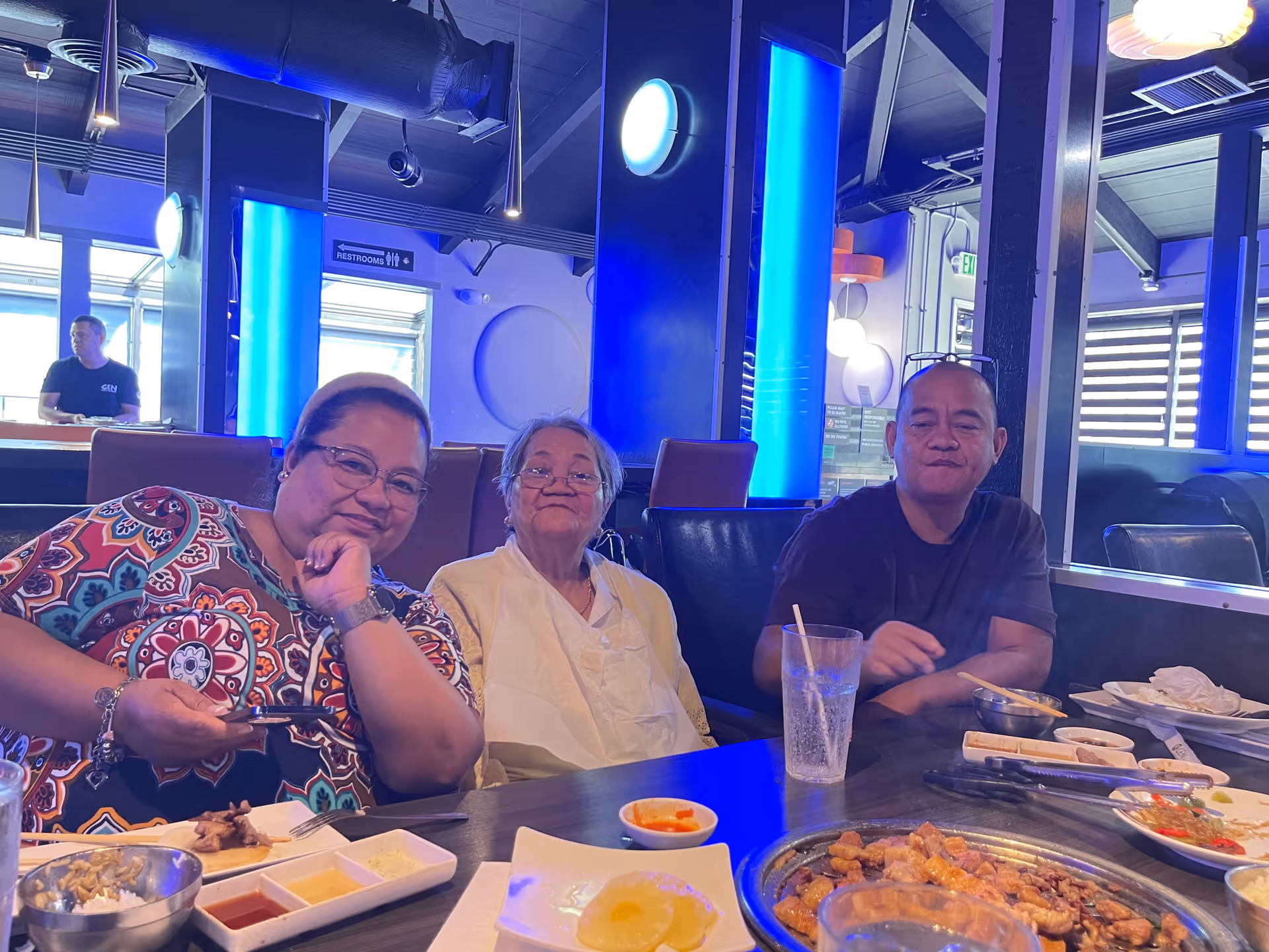 Three people sitting at a dining table in a restaurant with various dishes and drinks in front of them. The background features blue lighting and a sign indicating restrooms.