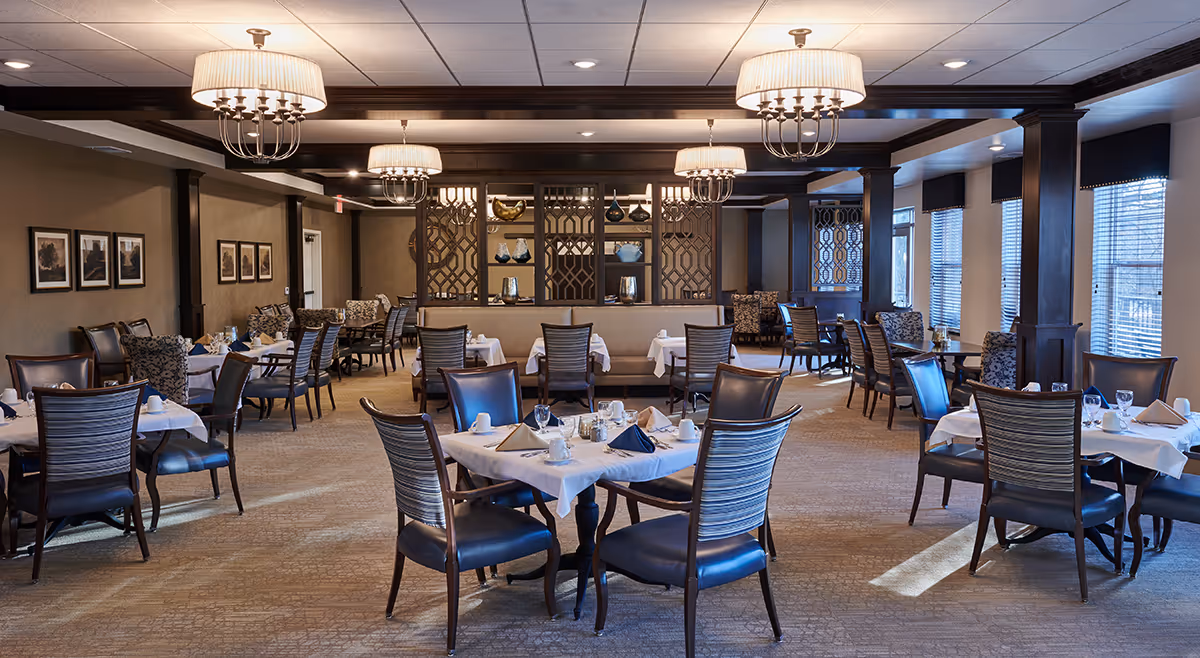 A spacious, well-lit dining room with multiple tables covered in white tablecloths, set with napkins, cups, and glasses. The room features dark wooden chairs with cushioned seats, decorative wall art, large windows with blinds, and elegant ceiling light fixtures.