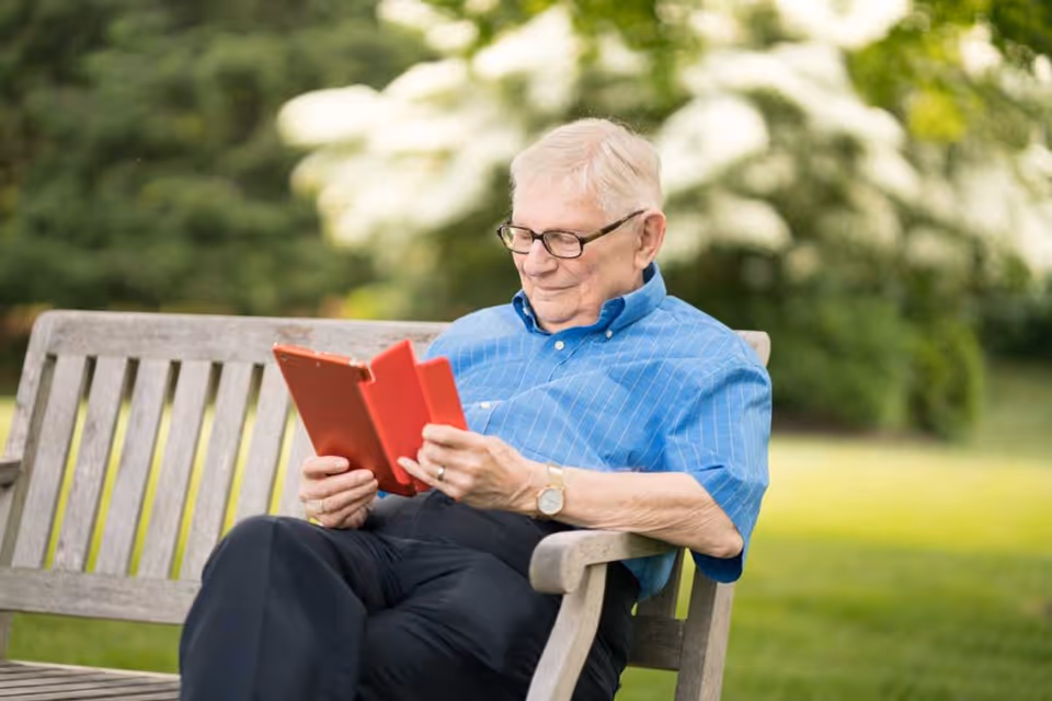An elderly man wearing glasses and a blue shirt is sitting on a wooden bench outdoors, reading a book with a red cover. The background is blurred with greenery and white flowers.