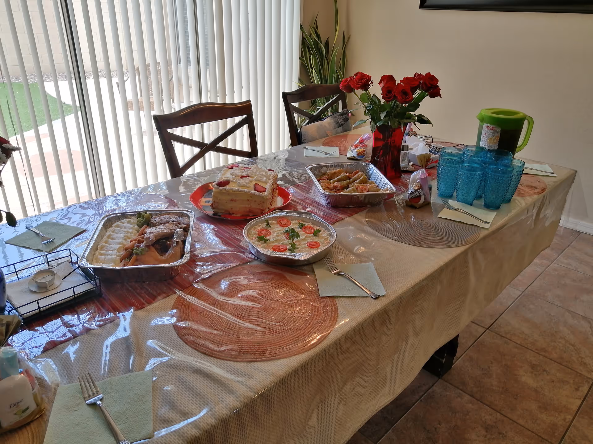 A dining table set for a meal with various dishes including a cake with strawberries, a tray of cooked meat with mashed potatoes, a bowl of salad with sliced tomatoes, and a tray of spring rolls. There is a red vase with red roses, a green pitcher, and several blue drinking glasses on the table. The table is covered with a beige tablecloth and a clear plastic cover. Four wooden chairs are around the table, and vertical blinds cover a large window in the background.