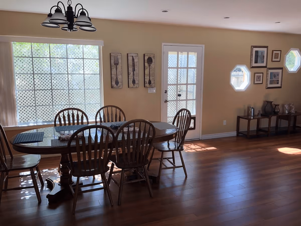 Sunlit dining area with a wooden table and six chairs on hardwood floors, a large window, and a glass-paneled door.