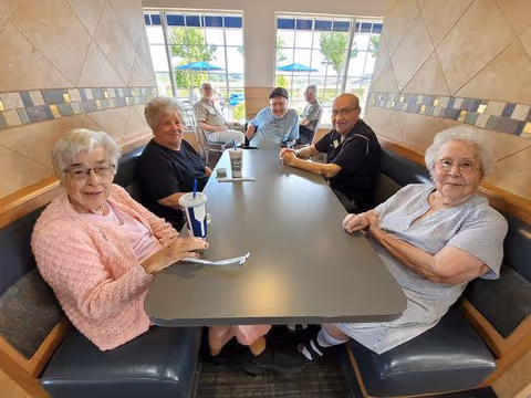 Six elderly people sitting around a rectangular table in a booth seating area inside a room with tiled walls and large windows showing an outdoor view with trees and umbrellas. They appear to be socializing and enjoying drinks.