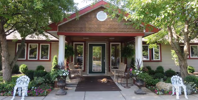 Front entrance of Heritage Manor of Lockport senior living community featuring a covered porch with white columns, outdoor seating, potted plants, and a cat resting on a brown mat. The building is surrounded by trees and landscaped flower beds.