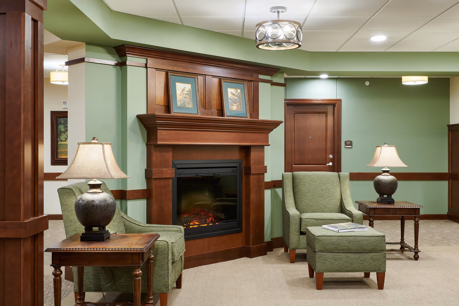 A cozy sitting area in a senior living facility with two green upholstered armchairs and a matching ottoman. Each chair is accompanied by a wooden side table with a lamp. The room features a wooden fireplace mantel with two framed bird pictures above it. The walls are painted green with wooden trim, and there is a wooden door labeled 323 in the background.