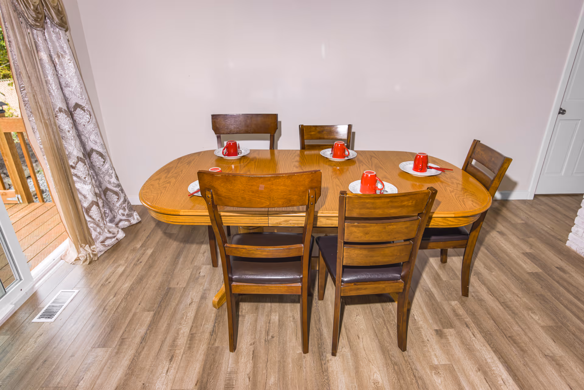 A wooden dining table set with six matching wooden chairs on a light wood floor. Each place setting has a white plate with a red cup and a red utensil. There is a sliding glass door with patterned curtains to the left, leading to an outdoor deck.