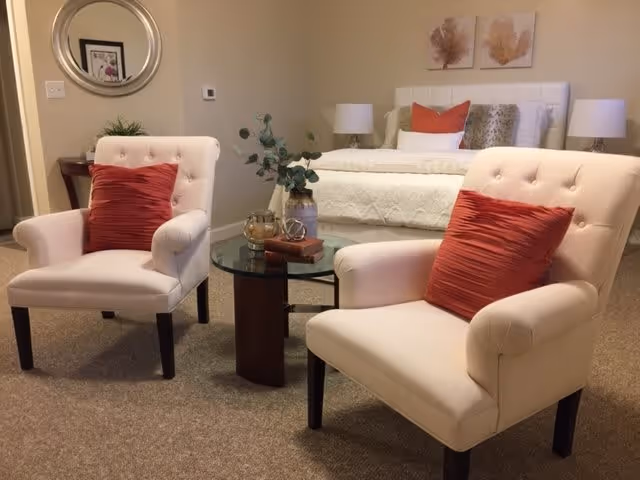 A cozy interior room featuring two cream-colored armchairs with red cushions facing a round glass coffee table. Behind the chairs is a neatly made bed with white bedding and decorative pillows, flanked by two bedside tables with lamps. The walls are decorated with framed artwork and a round mirror.