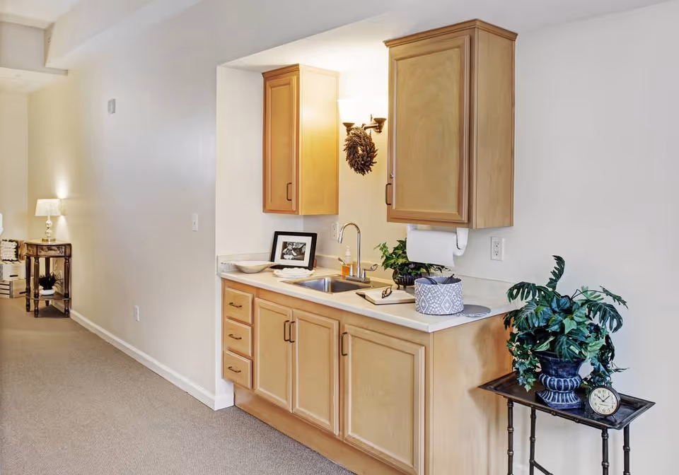 Interior view of a small kitchenette area with light wood cabinets, a sink, and countertop. There is a decorative wreath hanging on the wall above the sink, a framed photo, a bowl, and a small plant on the counter. To the right, a small table holds a potted plant and a clock. The background shows a hallway with a side table and a lamp.