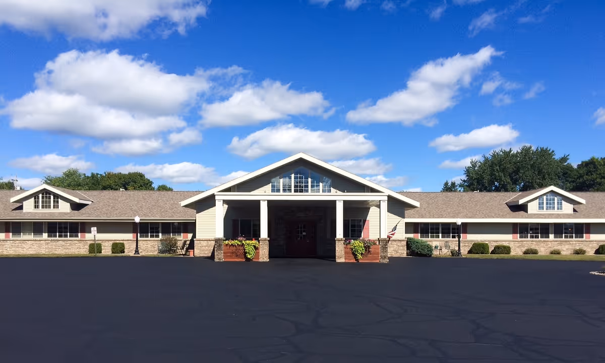 Front exterior view of a single-story senior living facility building with a large covered entrance, beige siding, brick accents, and multiple windows under a blue sky with scattered clouds.