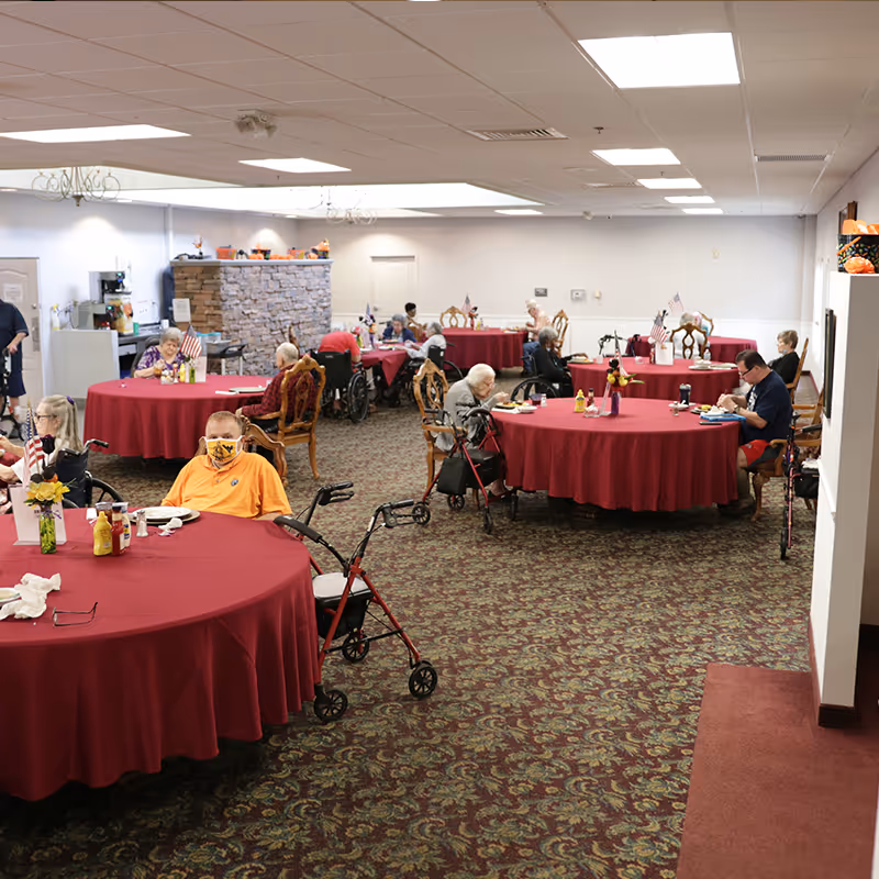 A dining room in a senior living facility with several round tables covered with red tablecloths. Elderly residents, some in wheelchairs and using walkers, are seated at the tables eating or engaging in activities. The room has patterned carpet, white walls, and ceiling lights. There is a stone accent wall with a counter area in the background.