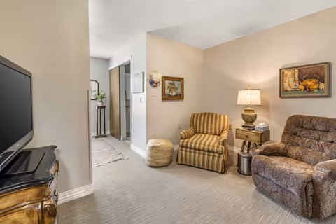 A cozy living room area with a striped armchair and a patterned recliner. A wooden side table with a lamp and a tissue box is placed between the chairs. The walls are decorated with framed paintings. A television is visible on a wooden stand to the left. The room has light-colored walls and carpeted flooring, with a hallway leading to another area in the background.