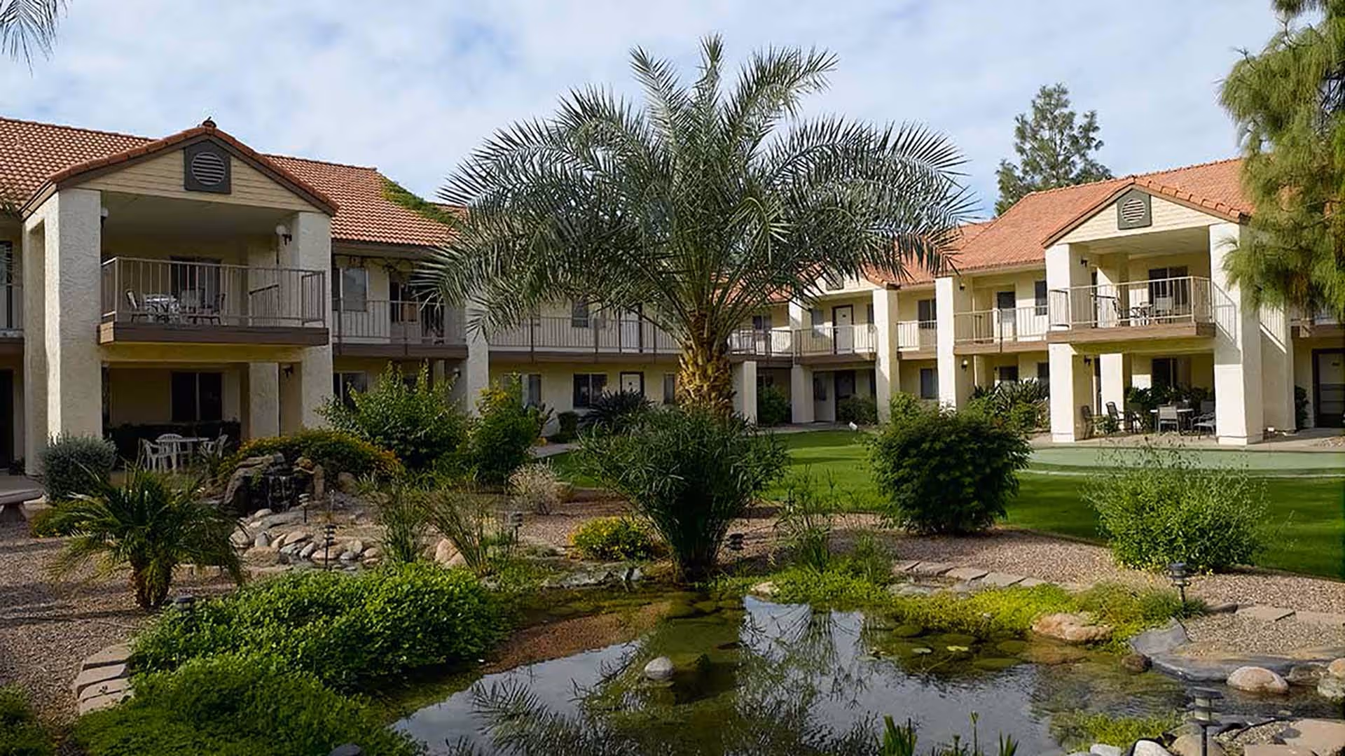 Outdoor courtyard area of a senior living facility with a small pond, various green plants, bushes, and palm trees. The two-story building with balconies and red-tiled roofs surrounds the courtyard.