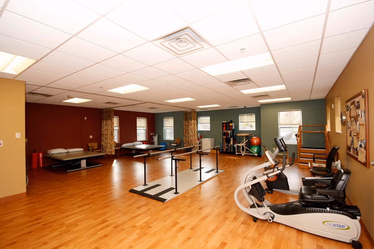A spacious rehabilitation or physical therapy room with wooden flooring, exercise equipment including a NuStep machine, parallel bars for walking exercises, therapy beds, exercise balls, and various therapy tools. The room has multiple windows with blinds, a bulletin board on the wall, and a ceiling with fluorescent lighting.
