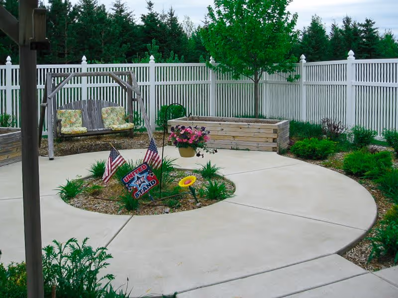 A peaceful outdoor garden area with a circular concrete pathway surrounding a small garden bed decorated with American flags and a 'United We Stand' sign. There is a wooden swing bench with floral cushions, a hanging flower pot, raised garden beds, and a white picket fence enclosing the space with trees and shrubs in the background.