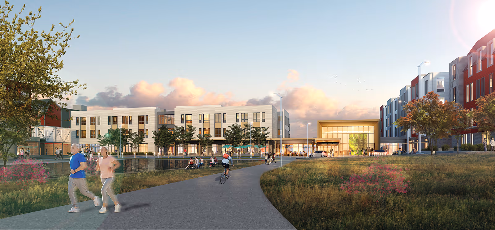 A modern senior living facility with multiple buildings surrounding a pond and landscaped green space. People are seen walking, jogging, cycling, and sitting on benches along a paved pathway. The sky is partly cloudy with a warm sunset glow.