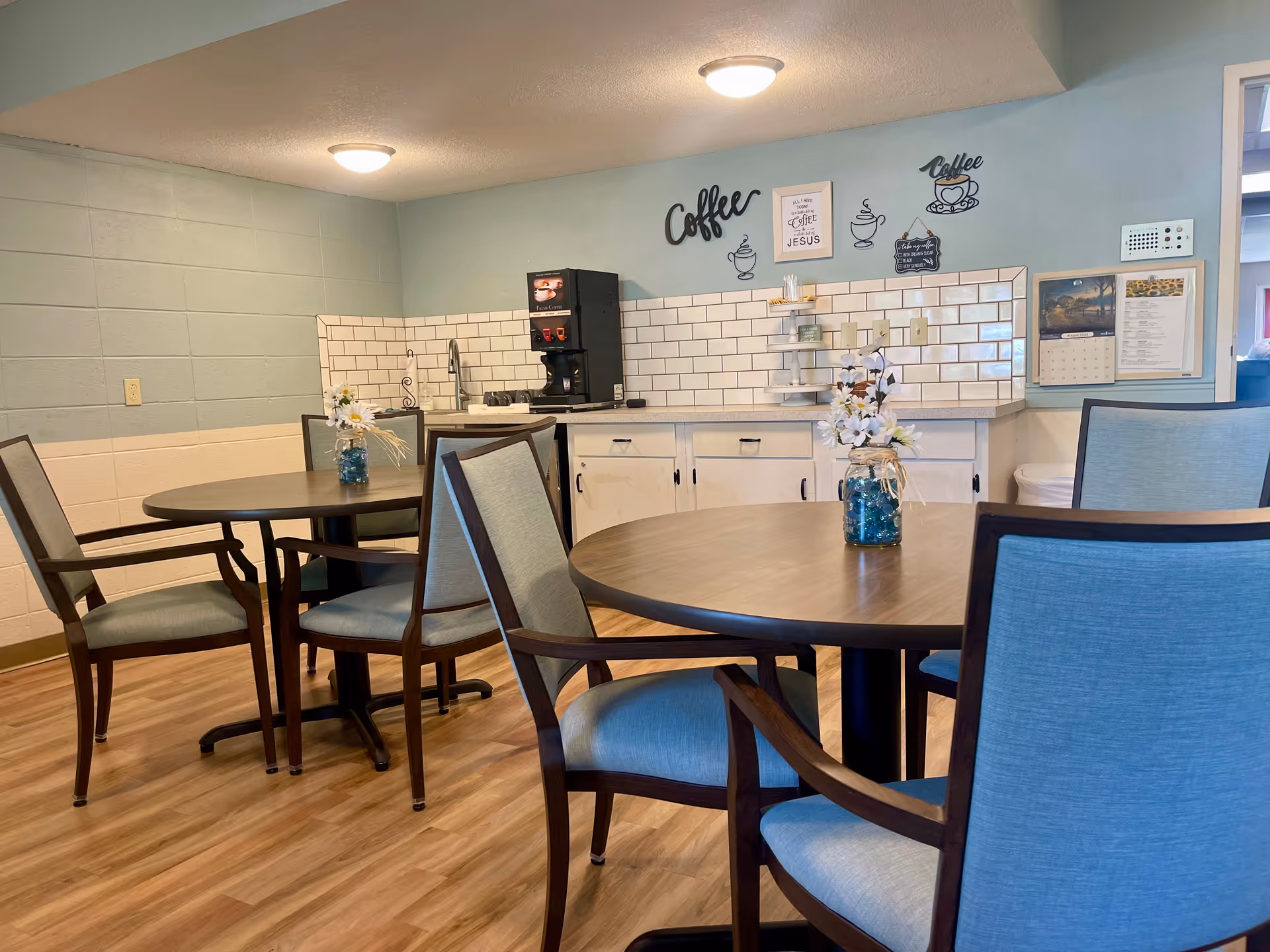 A cozy dining area with round wooden tables and blue cushioned chairs. Each table has a small vase with white flowers. In the background, there is a coffee station with a coffee machine, white tiled backsplash, and wall decorations related to coffee. The walls are painted light blue and light green, and the floor is wooden.