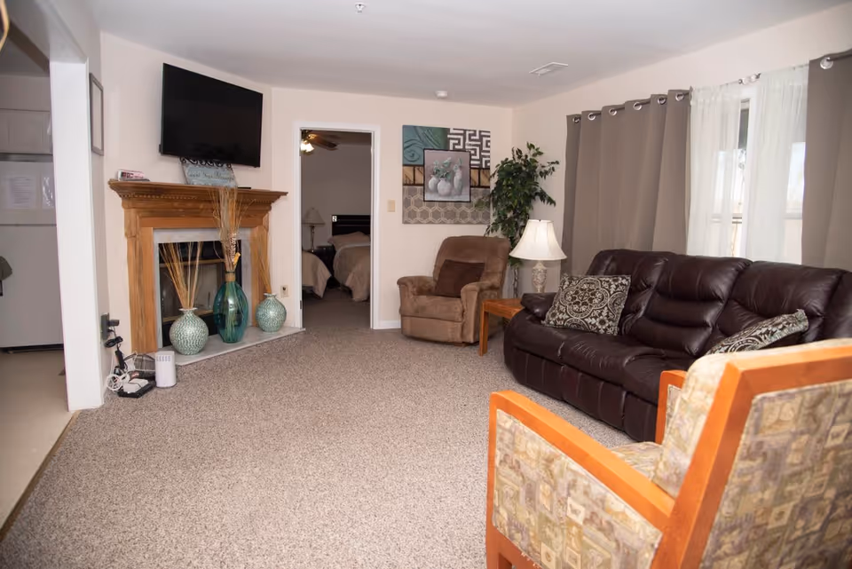 Cozy living room with a leather sofa, armchairs, TV mounted above a decorative fireplace, and a doorway leading to a bedroom.